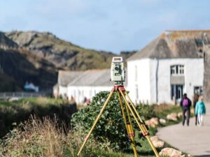 Surveying instrument on a tripod near a coastal path, with white-washed buildings in the background.