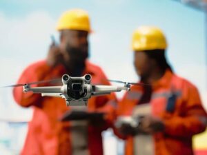 Close-up of a drone flying toward the camera with two construction workers in orange uniforms and yellow helmets in the background.